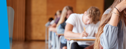 Learners in a hall sitting an exam, one learner with hand in head with a stressed experssion.